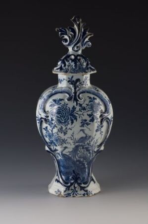  A traditional blue and white porcelain vase with a rounded body and ornate lid featuring intricate floral patterns and baroque-style embellishments against a gray background. Artist name and title are unknown.