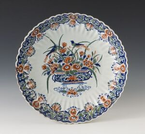  An ornate round ceramic plate with a scalloped edge, featuring a central floral motif in shades of deep cobalt blue and vibrant orange, surrounded by a detailed border pattern in matching colors on an off-white background. Artist name and title are unknown.