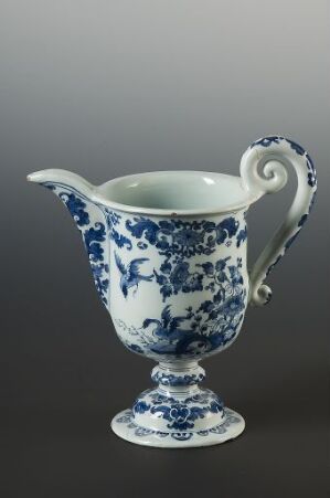  A traditional blue and white porcelain pitcher with intricate floral and landscape designs, standing on a pedestal foot with a gracefully curved handle, against a gray background.