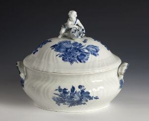  An oval-shaped porcelain tureen with a blue floral pattern, featuring ornate handles and a lid topped with a small white figure in a reclining pose against a neutral gray background. Artist name and title of the artwork are unknown.