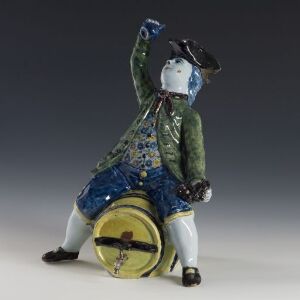  Ceramic figurine of a jovial man in an 18th-century-inspired green coat, light blue shirt, and vibrant blue pants with a gold band, playing a yellow drum while wearing a black tricorne hat.