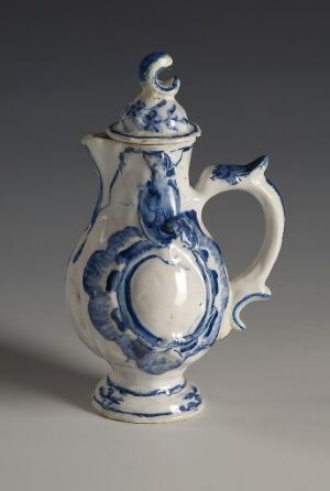  A white-glazed faience oil jug titled "Olie-Kande" with intricate hand-painted cobalt blue decorations. The jug has a bulbous body with a medallion-like blue motif in the center, an elegantly curved handle with blue accents, an ornate blue design along the spout, and a blue-painted lid knob. It is resting against a neutral grey background. Artist name is unknown.