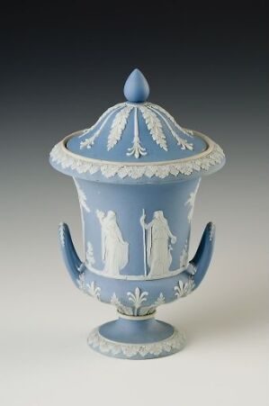  An ornate powder blue and white decorative urn with curved handles and a conical-lidded top, featuring classical white silhouettes within vertical striped panels and accented with a white foliage-inspired pattern.