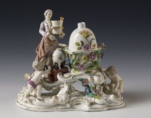  A detailed porcelain figural group featuring an ornate inkwell adorned with pastel flowers, with a female figurine in pale purple attire to the left, and a small dog to the right, all set on a baroque-style white base against a neutral grey background.