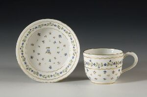  A porcelain teacup and saucer set on an off-white background, with delicate blue, green, and purple floral designs and blue trim on the edges. The saucer lies flat with the design visible inside while the cup is upright with a visible handle to the right, showcasing the floral pattern on its exterior.