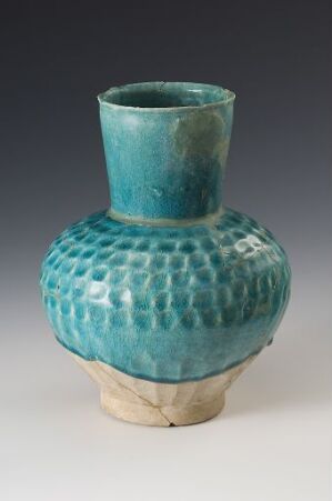  A ceramic vase with a bulbous body and narrow neck, featuring a textured honeycomb pattern glazed in shades of turquoise and sea green, sitting against a gray background. The base of the vase is unglazed, revealing the natural beige color of the clay. Artist name and title are unknown.