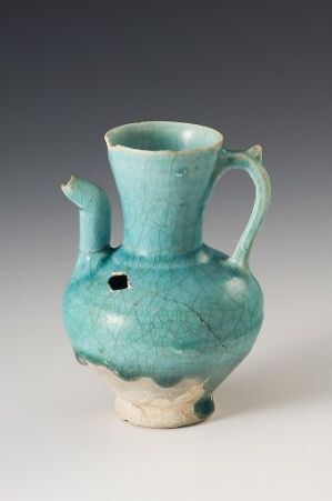  A turquoise-glazed ceramic pitcher with a single handle and a rounded body sitting against a neutral background. The pitcher has an antique finish with a crackled texture and an unglazed beige base.