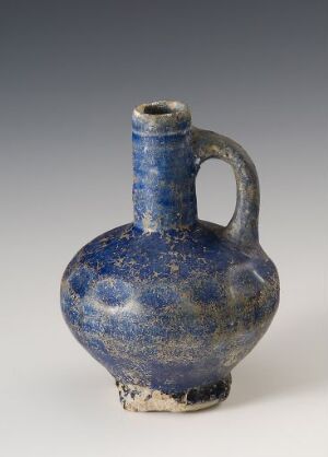  An ancient ceramic jug with a rounded body and single handle, featuring a deep blue glaze with hues of lighter blue and white speckles, and a bare clay base with earthly deposits.