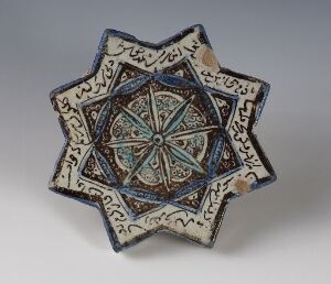  A star-shaped ceramic decorative object with a concave design and eight points, featuring a central teal blue star pattern surrounded by dark blue and orange accents, with Arabic script along the arms outlined in dark brown or black against a creamy white background.