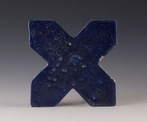  A textured deep blue sculptural object in the shape of the capital letter 'X', standing against a light grey background, with no artist name or title provided.