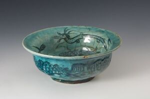  A historic Asian-style ceramic bowl with a glossy turquoise blue glaze, decorated with darker blue patterns of floral and mythological scenes inside and circular motifs outside, set against a neutral gray background.