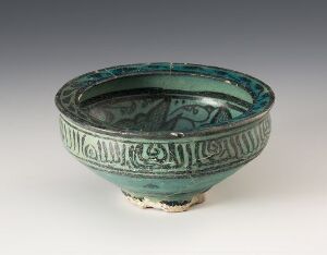  An ancient turquoise-glazed ceramic bowl with ornate geometric patterns, standing on three stubby feet against a neutral grey background. Artist and title unknown.