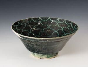  A handcrafted ceramic bowl with a dark teal glaze and fine, light green crazing on both the inside and outside, creating an intricate web-like pattern. The bowl narrows to a small rounded foot, and the image is taken against a neutral, softly textured background.