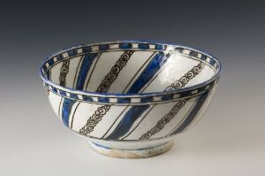  A ceramic bowl with a flared rim and rounded bottom, decorated with alternating vertical stripes of dark blue and patterned white with linked circle motifs. The design is complemented by a blue-bordered rim and the bowl sits against a gray background.