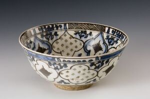  An intricately designed ceramic bowl with a blue and white pattern featuring stylized floral motifs and geometrical shapes, with a thin brown footed base. The title and artist are unknown.