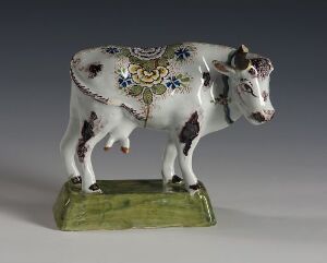  A ceramic cow figurine with white and brown patches stands on a green base; it features a blue, yellow, and green floral pattern with gold accents on its back.