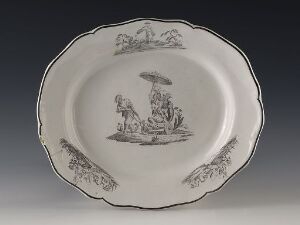  An oval-shaped ceramic plate with a monochromatic design, featuring a central scene of two figures beneath an umbrella and decorative foliage patterns around the edge. The entire plate is in shades of light and dark grey. Artist name and title are unknown.