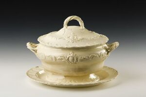  A cream-colored ceramic lidded tureen with embossed garlands and gilded accents, placed on a matching underplate against a light grey background.