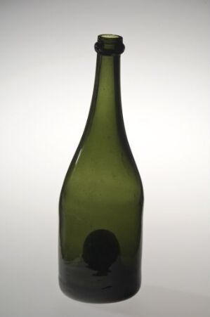  A tall slender green glass bottle with a rounded punt at the bottom stands against a background that graduates from white to light gray. The bottle has a simple traditional design with no labels or markings.