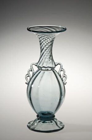  A transparent glass vessel with a bluish tint and decorative elements, featuring a bulbous lower body, intricate looping handles, and a wide, patterned upper brim against a neutral gray background. Artist name and title are unknown.