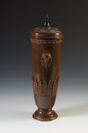  An elaborately carved cylindrical object, possibly wooden, with rich brown hues and a decorative gold band at the base, featuring intricate patterns and a shield-like emblem on the front, topped with a conical lid and a small finial.