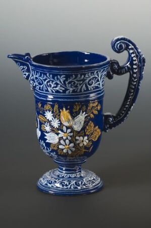  An ornate cobalt blue ceramic ewer with white and gold floral motifs and intricate scrollwork throughout, including on its elegantly curved handle and pedestal base, against a neutral background. Artist name and title are unknown.