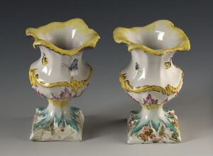  A pair of symmetrical porcelain vases with scalloped rims edged in yellow and gold, cobalt blue and gold acanthus-like scrollwork on the upper part of the body, pastoral scenes above the base, and a naturalistic rock-like base with painted green and purple foliage.