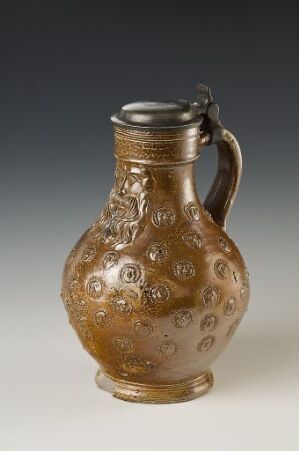  A translucent amber-colored glass pitcher with a metal lid and handle, adorned with raised circular designs with floral motifs, against a neutral-gray gradient background.