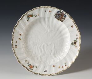  An antique scalloped-edge plate with an off-white base color, embellished with small, spaced-out floral patterns in soft shades of orange, green, brown, and blue along its rim, and a larger, colorful emblem resembling a coat of arms near the top. The plate displays a textured pattern in the center, suggestive of its historical nature, possibly from European origin.