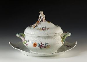  An antique porcelain soup tureen with floral patterns and leaf-shaped green handles on a matching oval underplate, featuring a decorative knob on the lid. The tureen's colors include shades of white, green, purple, pink, orange, and yellow, highlighting the detailed craftsmanship.