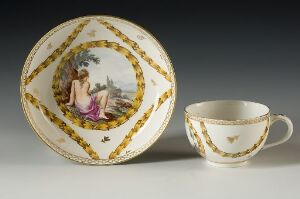  A porcelain tea set with a saucer featuring a central medallion with a classical scene of a lady in a purple robe and surrounded by a gilded chain design. The matching teacup has the same golden motif along the rim and a small star-shaped design inside.