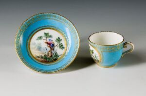  An antique porcelain tea set with a turquoise and golden bird-themed saucer and a matching tea cup, both decorated with gold accents and intricate designs.