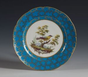  An antique decorative plate with a cobalt blue and gold-trimmed rim that surrounds a central white medallion depicting a colorful bird perched on a branch against a white background.