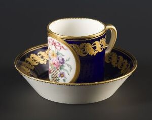  An antique cobalt blue and white porcelain tea cup with gold accents and a detailed floral design, paired with a matching saucer, against a neutral gray background.