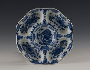  An ornate blue and white ceramic plate with a floral pattern and wavy edges, displayed against a neutral background.