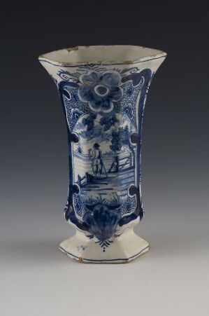  An antique hexagonal-shaped vase featuring intricate blue patterns and pastoral scenes on a glossy white porcelain background, indicative of traditional Delftware or Chinese blue and white pottery.