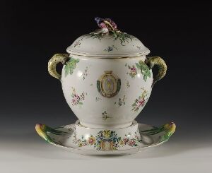  An ornate white porcelain soup tureen with a lid, featuring elaborate gold and green handles, a lifelike purple flower handle on the lid, and delicate floral patterns. It rests on a matching platter against a dark gray background.