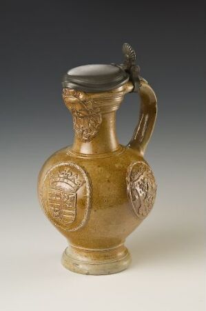  A small, antique amber glass jug with intricate embossed tree or floral medallions, featuring a curved handle and a flared neck, displayed against a gradient grey backdrop.