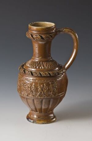  A photo of a brown stoneware pitcher titled "Bondedans" by Jan Emens Mennicken. The pitcher has a round body with finely detailed relief decoration depicting figures in a dance, a single curvy handle, and a narrow neck with a slightly flared opening. The piece exemplifies skilled turning and modeling techniques, finished with begitning, salt glaze, and pressed relief decoration. It stands against a neutral grey background that accentuates the rich brown tones and the glossy texture of the salt-glazed surface.