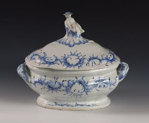  An oval-shaped historic porcelain tureen with intricate blue patterns on a white background, featuring scroll-like handles and a small bird figure atop the lid as a finial.