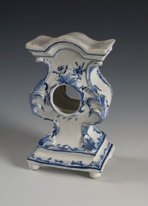  A decorative faience clock, lacking its mechanism, with a glossy white surface and hand-painted cobalt blue floral designs. The clock features a curvy, asymmetrical shape, indicative of a refined decorative style. Artist unknown.