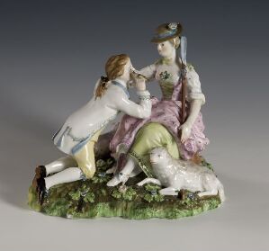 A porcelain figurine titled "Hyrdegruppe," featuring a male and a female figure in 18th-century pastoral dress, with the man kneeling and holding the woman's hand as she sits with a sheep in her lap, all set upon a grassy base with intricate detailing.