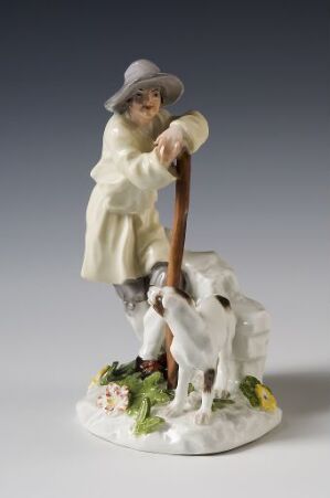  A porcelain figurine of a young shepherd boy wearing a cream coat and a lavender-grey hat, leaning on a brown stick, with a white and black dog beside him on a base with painted grass and flowers.