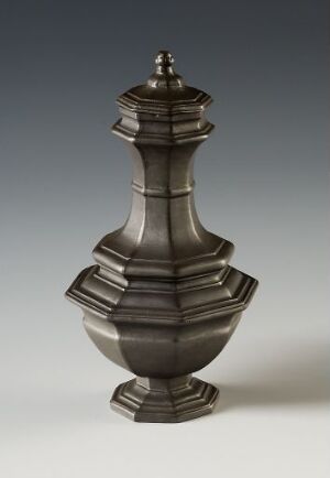  An antique dark gray metallic vessel with a symmetrical design featuring multiple bulbous sections, a narrow neck, and a finial-topped lid, set against a plain, light background.