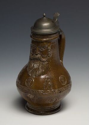  An antique brown jug with a metal lid, featuring a relief design of a face and intricate patterns reminiscent of historical craftsmanship.