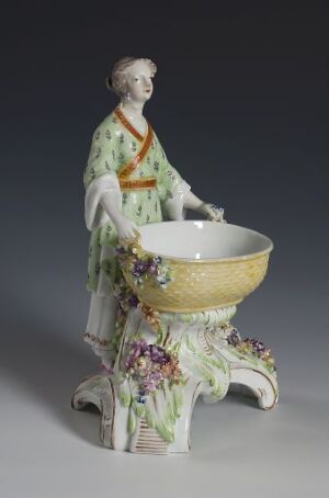  Porcelain figurine of an 18th-century woman in a pale green and polka-dotted dress with a gold bowl and an ornate floral base, displaying intricate details and soft pastel colors.
