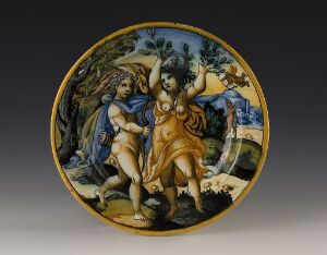  A glazed faience plate titled "Apollo og Daphne" by an unknown artist, showcasing a hand-painted mythological scene with vibrant colors. The plate features figures Apollo and Daphne, with Daphne depicted in mid-transformation into a tree, set against a landscape backdrop, framed by a dark yellow border.