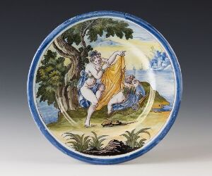  A round fajanse plate featuring the classical mythological scene of Venus and Amor (Cupid), with Venus semi-nude draped in a golden garment, and Cupid with outstretched wings, set against a pastoral backdrop with blue sky and green foliage, encircled by a cobalt blue rim.