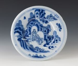  A round blue and white porcelain plate with a central figure surrounded by intricate flora and stylized patterns, positioned against a soft gray background.