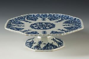  A pedestal cake stand with a classic blue and white design, featuring a scalloped rim and intricate blue floral and geometric patterns against a white background, evoking a vintage, traditional style.